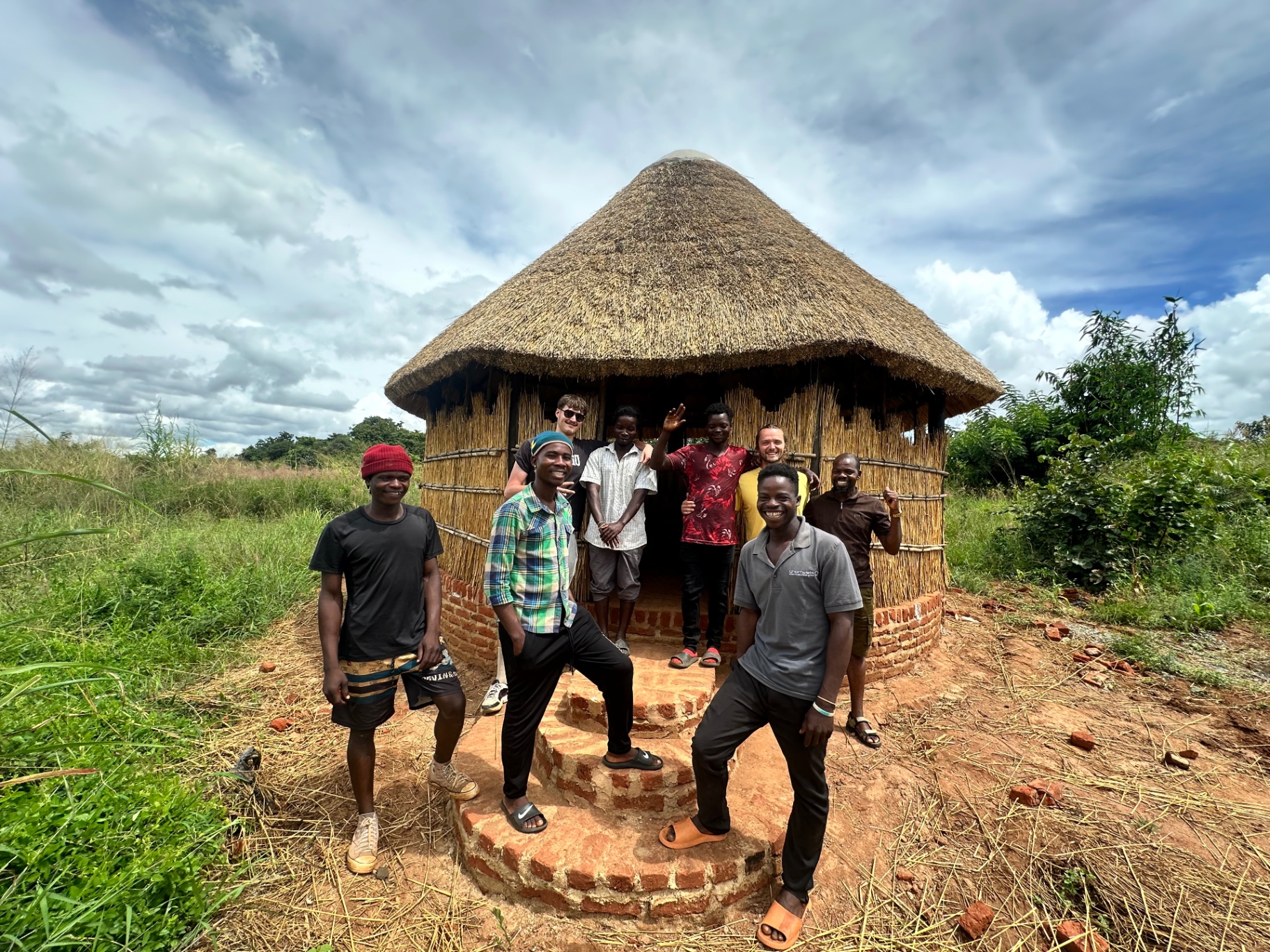 A smiling team standing in front of a newly built thatched round house on the land