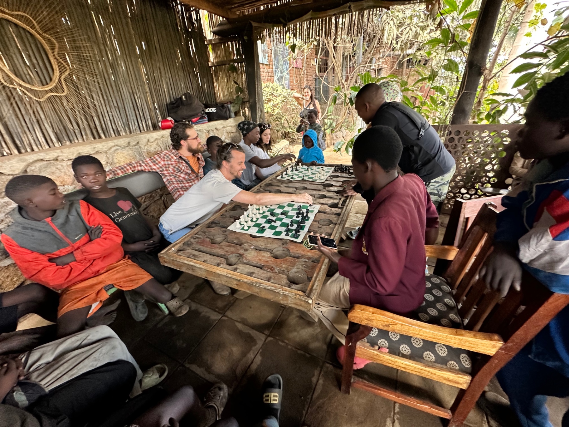 Children and adults playing chess together at a long wooden table under a shaded shelter